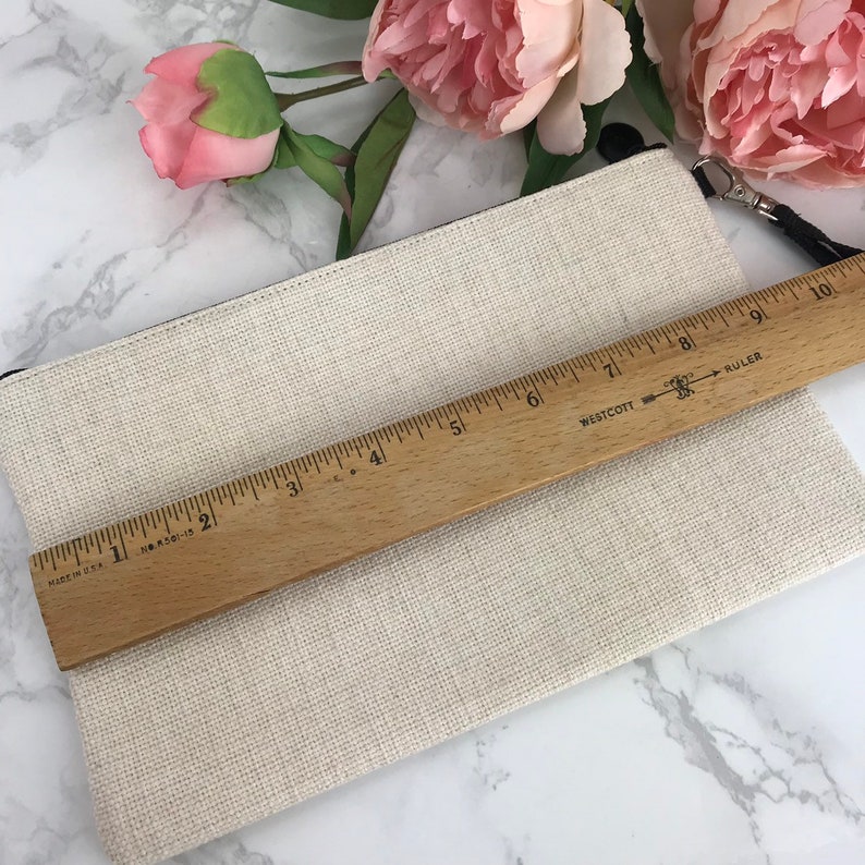 May include: A beige canvas zippered pouch with a black zipper pull and a black drawstring. The pouch is being measured with a wooden ruler that reads "Westcott Ruler".