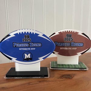 May include: Two football-shaped trophies with a blue and brown design. The trophies have a white base and a black base. The trophies have the text "9U 2025 Perseus Kouns Offensive MVP" on them. The blue trophy has a blue and white "M" logo on it.