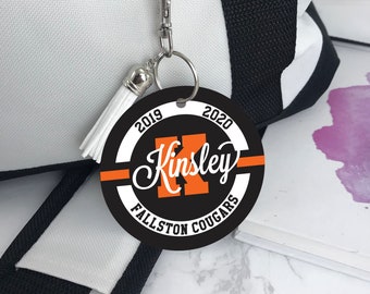 May include: Black and white round keychain with a white border and orange text. The text reads "2019 Kinsley 2020 Fallston Cougars".