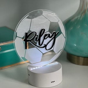 May include: A white acrylic night light shaped like a soccer ball with the name "Riley" written in black cursive. The light has a white base with a small button on the side and three black dots on the bottom.