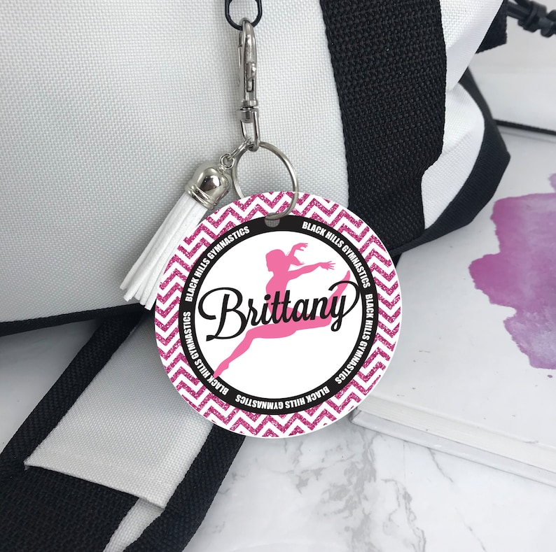 May include: Pink and black round tag with a white tassel. The tag has a pink glitter background with a chevron pattern. The tag says "Black Hills Gymnastics" around the edge and has a silhouette of a gymnast in the center. The name "Brittany" is written in black cursive above the gymnast.