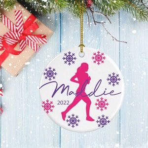 May include: White ceramic Christmas ornament with a pink silhouette of a runner and the name "Maddie" in purple script. The ornament is decorated with purple and pink snowflakes and the year "2022" in purple.