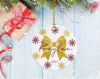 May include: White ceramic Christmas ornament with a gold bow and the name "Kaylee" written in red cursive. The ornament is decorated with gold and red snowflakes and the year "2022" is printed at the bottom.