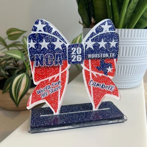 May include: A decorative bow-shaped trophy with red, white, and blue glitter accents. The bow features the text "NCA 2026 Houston TX" and "World Cup All Stars Campbell". The trophy sits on a rectangular base.