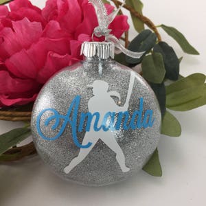 May include: Glitter-filled clear glass ornament with a silver cap and a silver ribbon. The ornament features a white silhouette of a softball player and the name "Amanda" in blue script. The ornament is surrounded by pink flowers and green leaves.