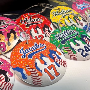 May include: A collection of personalized baseball-themed tags with names and numbers. Each tag features a baseball design with colorful dripped icing and sprinkles. The names are in a script font, and the numbers are in a block font.