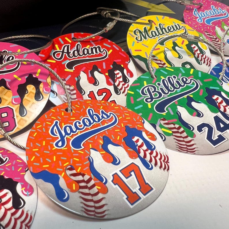 May include: A collection of personalized baseball-themed tags with names and numbers. Each tag features a baseball design with colorful dripped icing and sprinkles. The names are in a script font, and the numbers are in a block font.