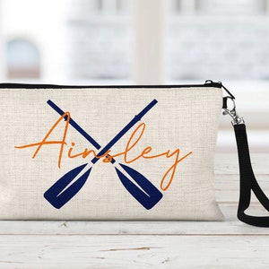 May include: A white canvas makeup bag with a black zipper and a black strap. The bag has a design of two crossed oars in navy blue and the name "Ainsley" in orange.