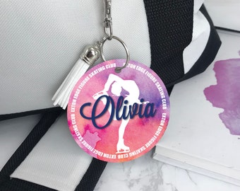 May include: A round, pink and purple watercolor-style keychain with a silhouette of a figure skater. The keychain has the text "EXTON EDGE FIGURE SKATING CLUB" repeated around the edge and the name "Olivia" in a navy blue font.