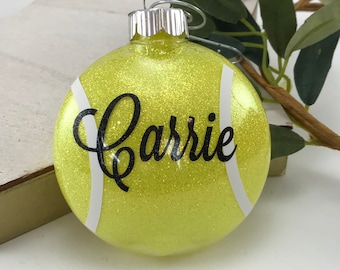 May include: A yellow glittered glass Christmas ornament with the name "Carrie" in black script. The ornament has white stripes and a silver metal cap with a wire loop for hanging. The ornament is designed to resemble a tennis ball.