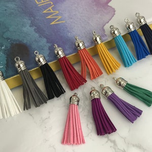 May include: A collection of 13 colorful suede tassel keychains with silver metal tops. The tassels are in various colors, including white, gray, black, red, orange, yellow, green, blue, and purple.