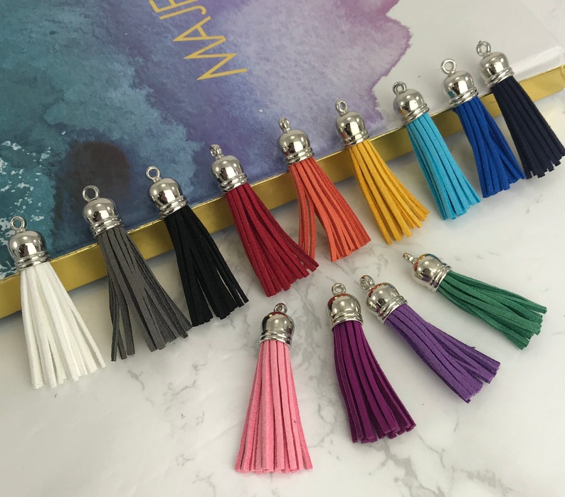 May include: A collection of 13 colorful suede tassel keychains with silver metal tops. The tassels are in various colors, including white, gray, black, red, orange, yellow, green, blue, and purple.