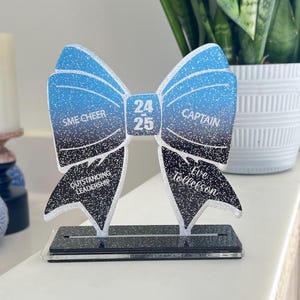 May include: A blue and black glitter bow award with the text "SME Cheer", "24-25", "Captain", "Outstanding Leadership", "Eve Tollefson" and a black base.