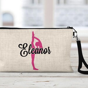 May include: A beige canvas makeup bag with a black zipper and a black strap. The bag has a pink silhouette of a gymnast in a backbend pose and the name "Eleanor" in black cursive lettering.