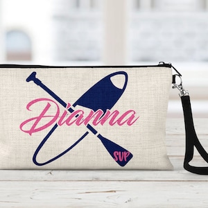 May include: A white canvas makeup bag with a black zipper and a black strap. The bag has a blue and white graphic of a paddleboard and paddle with the name "Dianna" in pink lettering and the word "SUP" in blue lettering.
