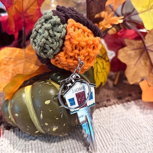 May include: A metal house-shaped keychain with a silver finish, blue windows, and a red door, featuring the word HOME printed in black. The keychain is attached to a silver key and a multi-colored crocheted topper in shades of brown, orange, and green.
