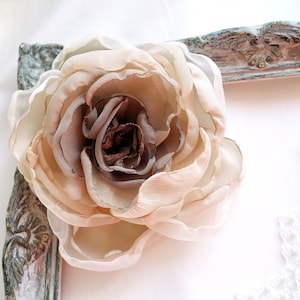 May include: A large, beige fabric flower with brown accents. The flower is made of multiple layers of fabric and has a delicate, ruffled appearance.