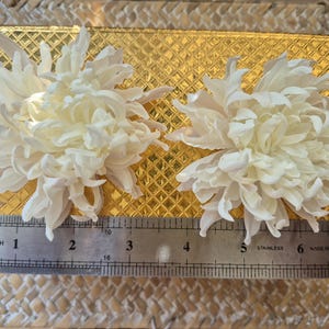 以下が含まれることがあります： 層状で尖った花びらを持つ2つの白い菊のような花が、ダイヤモンドのテクスチャが施された金色の長方形の箱の上に置かれています。ステンレス製の定規が手前にあり、花の幅が約14cmであることを示しています。