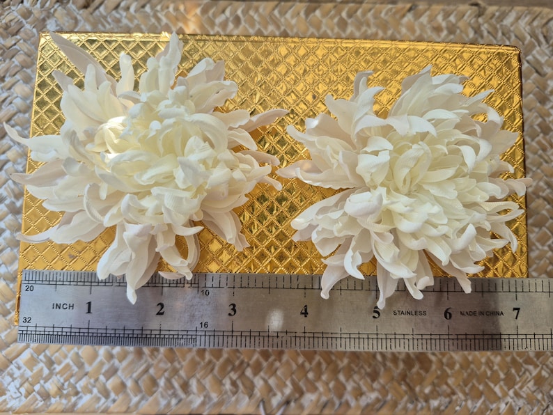 以下が含まれることがあります： 層状で尖った花びらを持つ2つの白い菊のような花が、ダイヤモンドのテクスチャが施された金色の長方形の箱の上に置かれています。ステンレス製の定規が手前にあり、花の幅が約14cmであることを示しています。