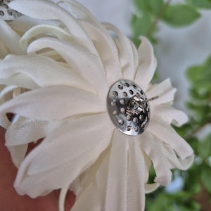 以下が含まれることがあります： オフホワイトの布製フラワーピアス。複数の花びらが重なったデザインです。各ピアスには、穴あき加工が施された円形のシルバーカラーの金属製バックプレートと、ピアスホール用の標準的なポスト留め具が付いています。