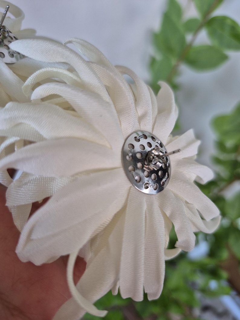 以下が含まれることがあります： オフホワイトの布製フラワーピアス。複数の花びらが重なったデザインです。各ピアスには、穴あき加工が施された円形のシルバーカラーの金属製バックプレートと、ピアスホール用の標準的なポスト留め具が付いています。