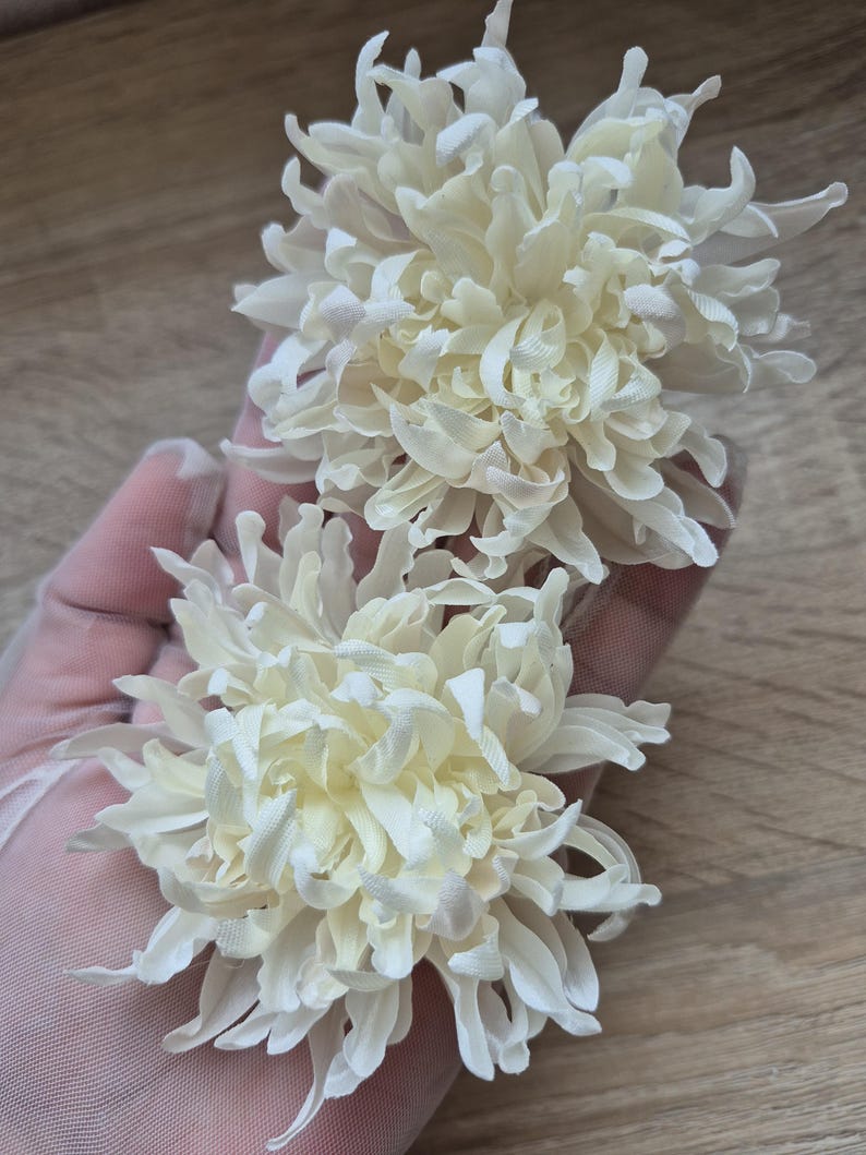 以下が含まれることがあります： 層状に重なりカールした花びらを持つ、オフホワイトの布製菊の花2輪。それぞれの花は、中心から放射状に広がる細く尖った花びらが密集した丸みを帯びた形状をしており、ボリューム感のある質感に仕上がっています。