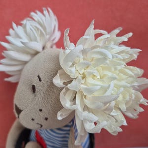 以下が含まれることがあります： 薄茶色の布製のぬいぐるみで、頭の両側に大きな白い菊の花が2つ付いています。濃い茶色の刺繍で目、鼻、口が表現されており、青と白のストライプ柄のシャツを着ています。