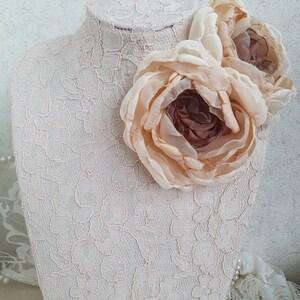 May include: Two large, pale peach-colored fabric flowers with a brown center, displayed on a white lace-covered mannequin bust.
