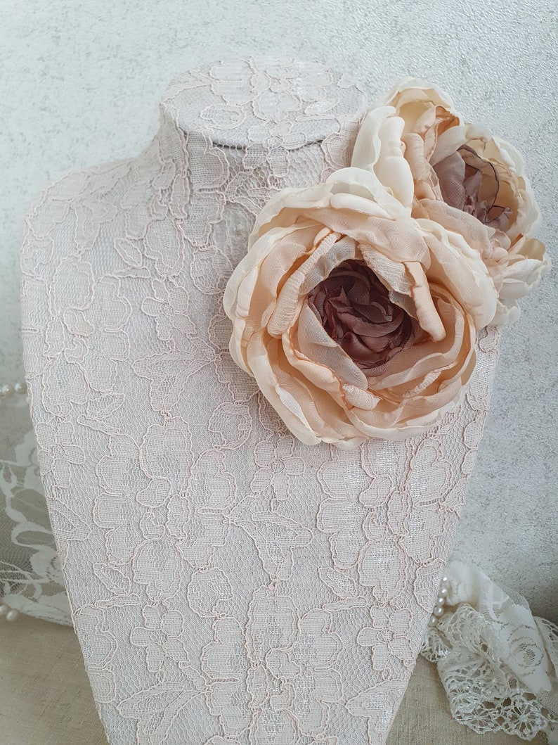 May include: Two large, pale peach-colored fabric flowers with a brown center, displayed on a white lace-covered mannequin bust.