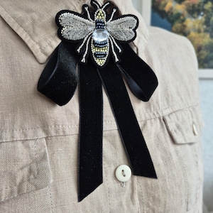 May include: A black velvet bow tie with a beaded bee brooch. The bee is embroidered with silver, gold, and black beads and has a clear rhinestone in the centre.