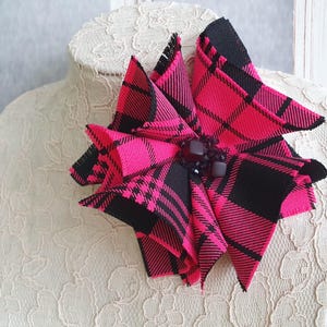 Puede incluir: Un broche de flor de tela a cuadros rosa y negro con un centro de pedrería negra.