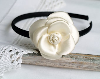 May include: A black headband featuring a large, off-white satin fabric flower attached to the side. The flower has multiple layered, rounded petals arranged in a circular, blooming pattern with a tightly rolled center, creating a smooth finish.