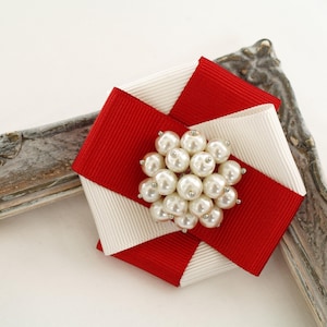 Könnte beinhalten: Dekorative Brosche mit einem geometrischen Design aus roten und weißen Bändern. Die Mitte der Brosche ist mit einer Ansammlung von Kunstperlen verziert. Die Brosche befindet sich vor einem weißen Hintergrund und einem dekorativen Rahmen.