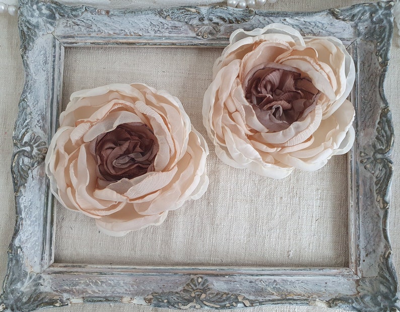 May include: Two large, pale pink fabric flowers with a brown center. The flowers are displayed in a white, ornate picture frame.