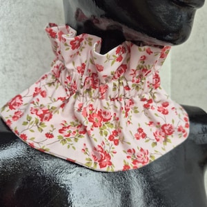 May include: A light pink fabric neck ruff featuring a repeating floral pattern of red roses and green leaves. The accessory has a gathered, ruffled collar design that sits around the neck, constructed with a soft, pleated fabric texture.