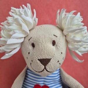 以下が含まれることがあります： ベージュの毛並み、茶色の刺繍の目、ダークブラウンの鼻を持つ動物のぬいぐるみ。花のような白い布を重ねた大きな耳が特徴で、青と白の横縞模様のシャツを着ており、小さな赤いハートのディテールがあしらわれています。
