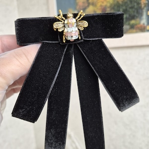 May include: A black velvet bow tie featuring a gold-tone bee brooch. The bee is decorated with small, colourful gemstones. The bow tie has long, rectangular tails.