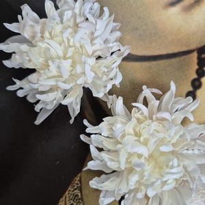 Puede incluir: Dos flores de tela blanca con numerosos pétalos finos y superpuestos dispuestos en una forma esférica densa. Los pétalos tienen un ligero brillo y están agrupados para crear una apariencia completa y texturizada, similar a un crisantemo.