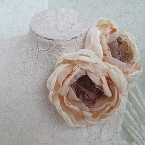 May include: Two large, pale peach-colored fabric flowers with a brown center, arranged on a white lace background.