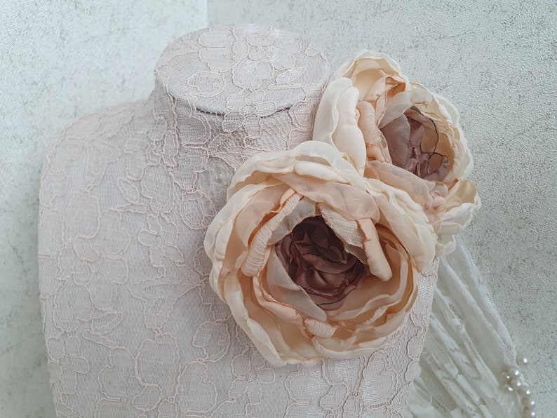 May include: Two large, pale peach-colored fabric flowers with a brown center, arranged on a white lace background.