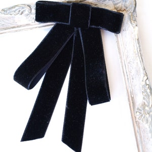 May include: A black velvet bow tie accessory featuring a central knot and two long, flat, hanging ribbon tails. The bow is constructed from a soft, dark fabric with a matte texture, designed to be worn as a decorative neck piece or brooch.