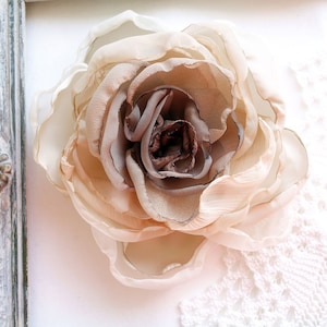 May include: A delicate, beige and brown fabric rose flower, with a ruffled texture, is displayed on a white background with a lace overlay.
