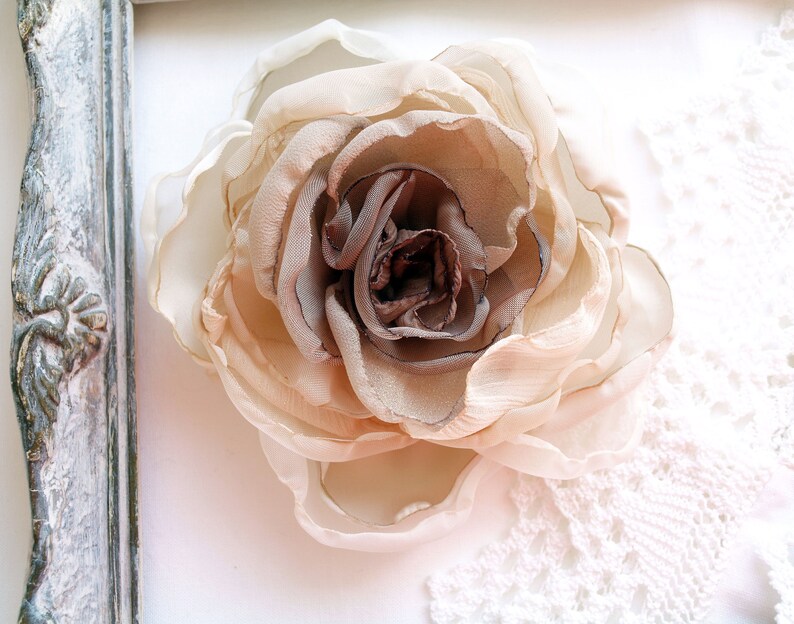 May include: A delicate, beige and brown fabric rose flower, with a ruffled texture, is displayed on a white background with a lace overlay.