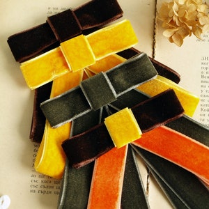 Puede incluir: Un conjunto de cuatro lazos de cinta de terciopelo en varios colores, incluyendo marrón, amarillo, naranja y verde. Los lazos están dispuestos en una pila, con el lazo más grande en la parte inferior y el lazo más pequeño en la parte superior.