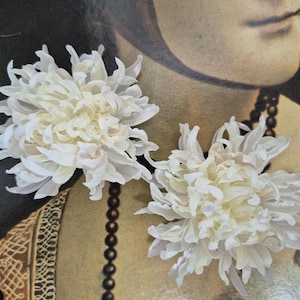 以下が含まれることがあります： 層状の尖った花びらを持つ2つの白い菊の花のヘアアクセサリー。花は密度が高く丸みを帯びた形状で、中心から放射状に広がる繊細で湾曲した花びらが特徴で、ヘアスタイリングに適したボリューム感と質感のある外観を作り出しています。