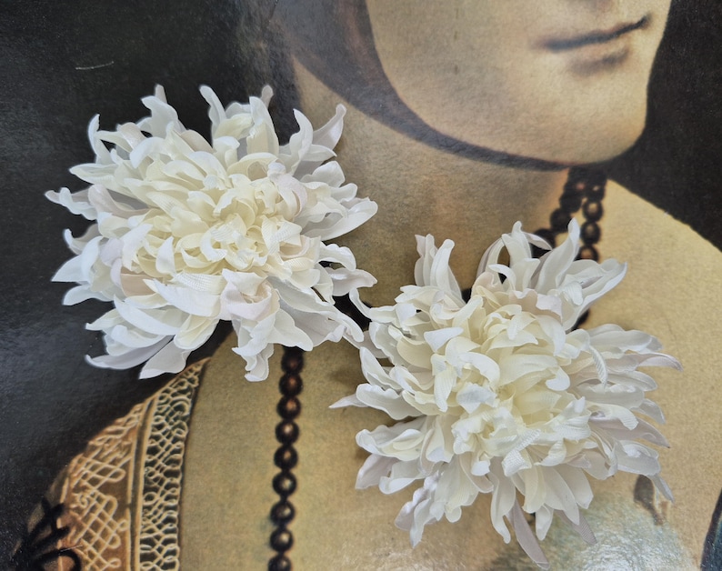 以下が含まれることがあります： 層状の尖った花びらを持つ2つの白い菊の花のヘアアクセサリー。花は密度が高く丸みを帯びた形状で、中心から放射状に広がる繊細で湾曲した花びらが特徴で、ヘアスタイリングに適したボリューム感と質感のある外観を作り出しています。