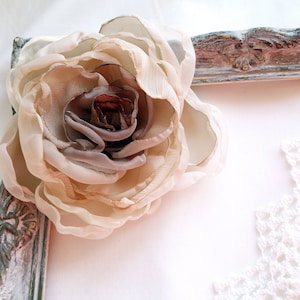 May include: A large, beige fabric flower with a brown center is placed on a white surface next to a distressed, white-painted picture frame.