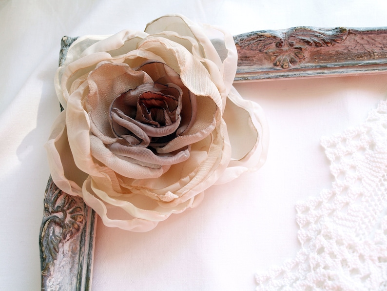 May include: A large, beige fabric flower with a brown center is placed on a white surface next to a distressed, white-painted picture frame.