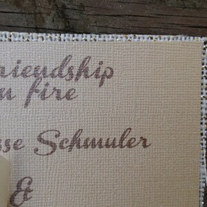 Puede incluir: Una tarjeta rectangular de color beige con superficie texturizada y borde de tela con hilos dorados. La tarjeta presenta texto impreso en marr&oacute;n que dice friendship, t on fire, Jesse Schmuler y un s&iacute;mbolo ampersand, con una cinta crema.
