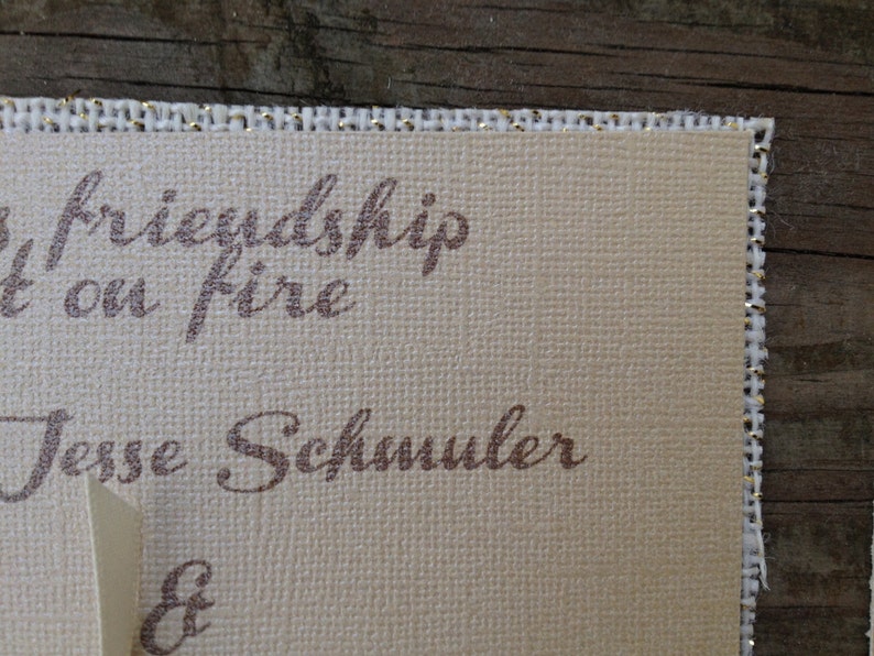 Puede incluir: Una tarjeta rectangular de color beige con superficie texturizada y borde de tela con hilos dorados. La tarjeta presenta texto impreso en marr&oacute;n que dice friendship, t on fire, Jesse Schmuler y un s&iacute;mbolo ampersand, con una cinta crema.
